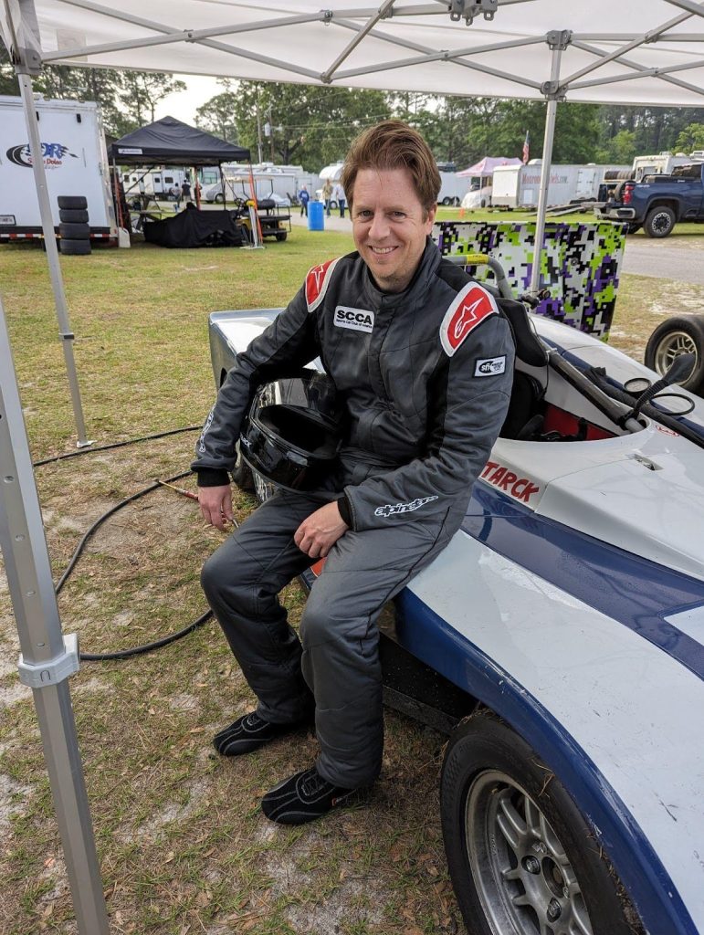 Driver in a race suit sitting on the nose of a Spec Racer Ford in the paddock
