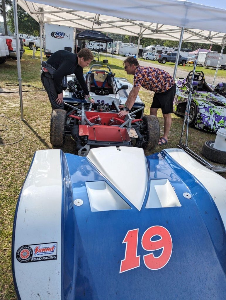 Two crew members working on the open front of Spec Racer Ford number 19 under a canopy