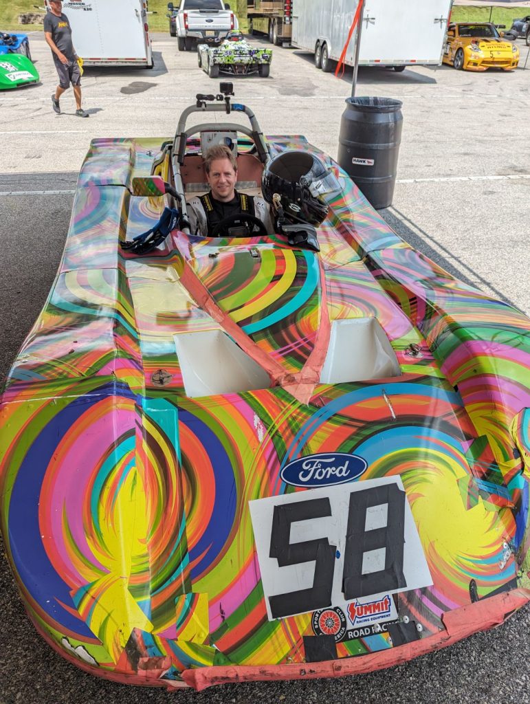 Driver seated in a colorfully wrapped Spec Racer Ford number 58 in the paddock
